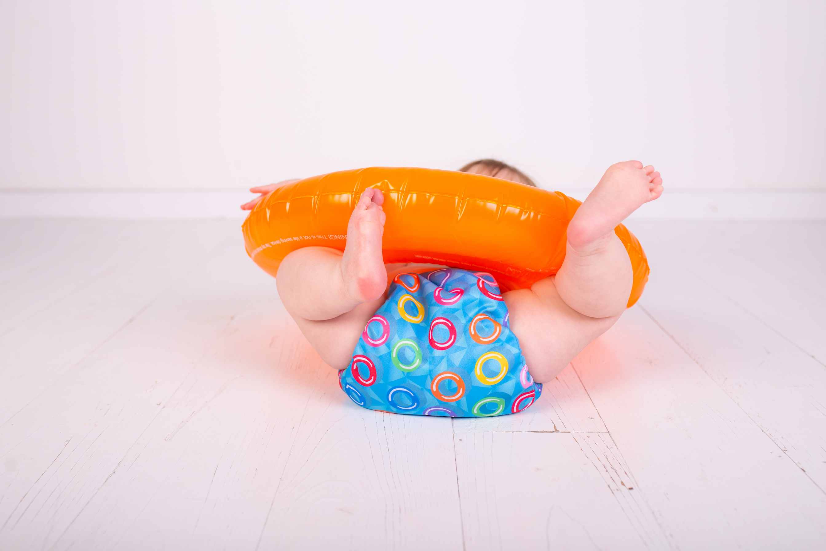 Child rolling on floor wearing TotsBots reusable washable swim nappy playing with orange rubber ring